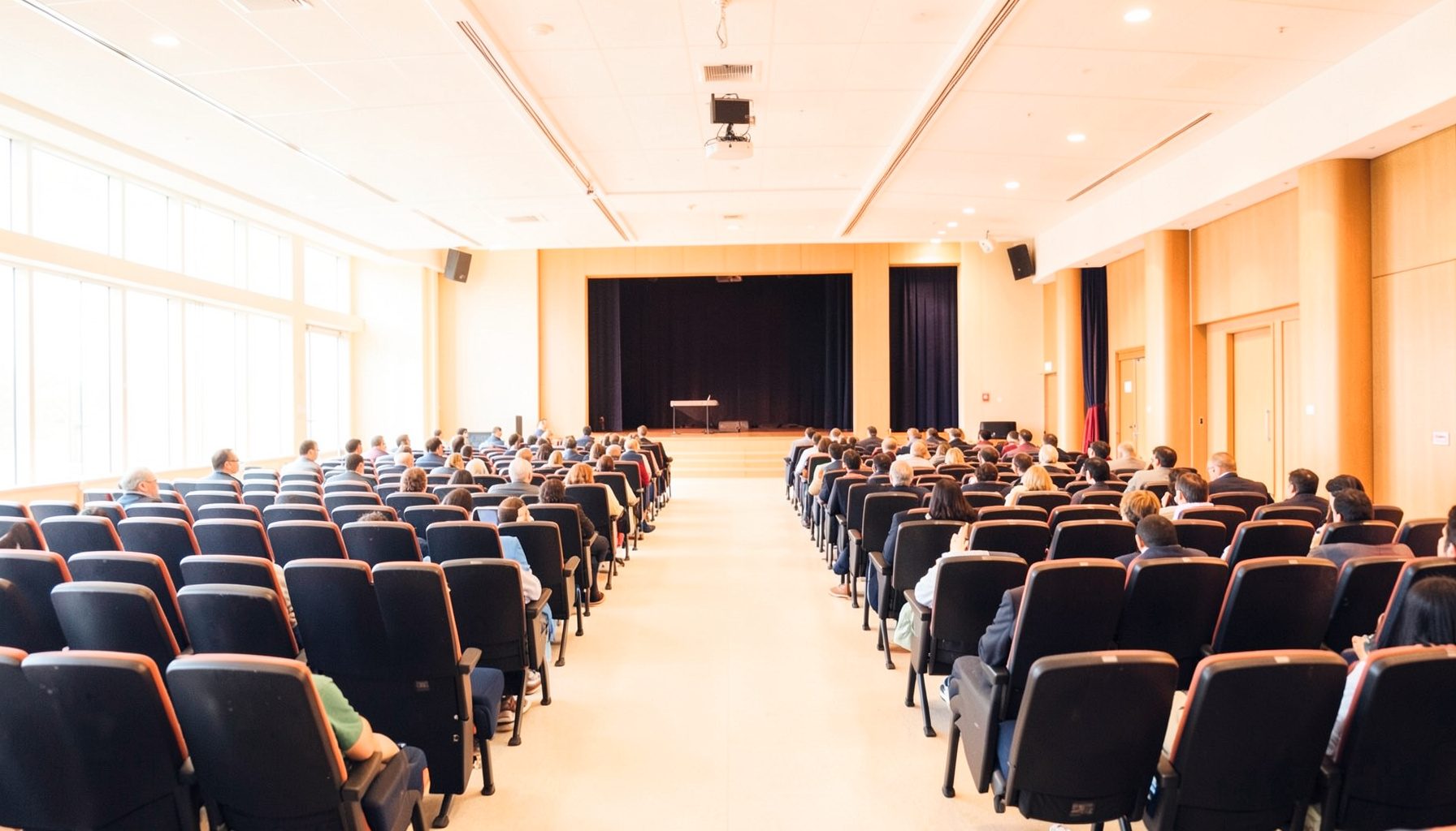 Wide-angle photograph of a bright conference hall with rows of seating and a stage, warm amber and deep navy tones, airy atmosphere