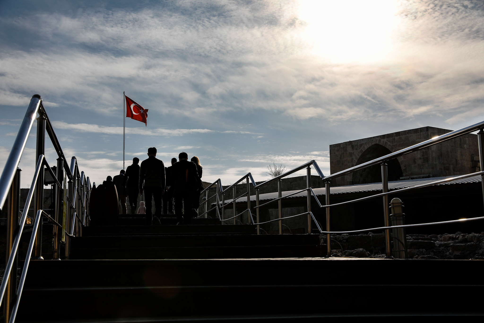 Silhouetted group of people walking up outdoor stairs toward a flag against a bright cloudy sky