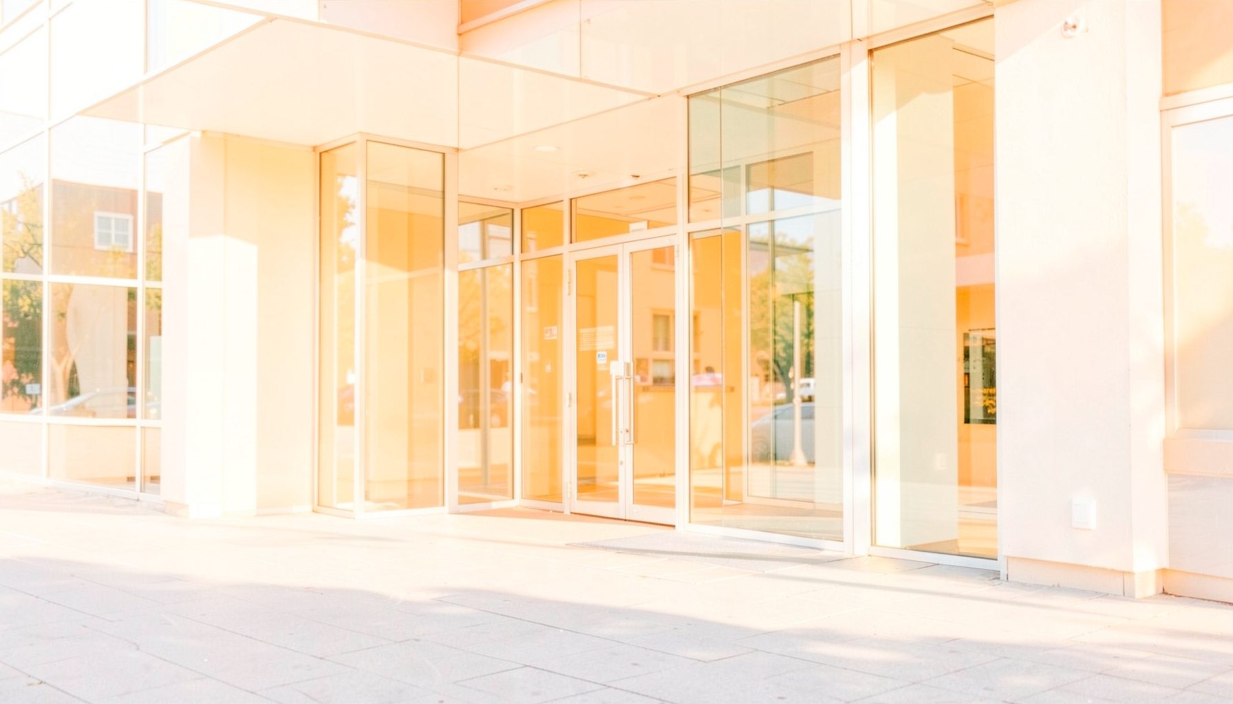 Bright, airy photograph of a modern municipal building entrance with glass doors and warm sunlight casting soft shadows across a welcoming public plaza