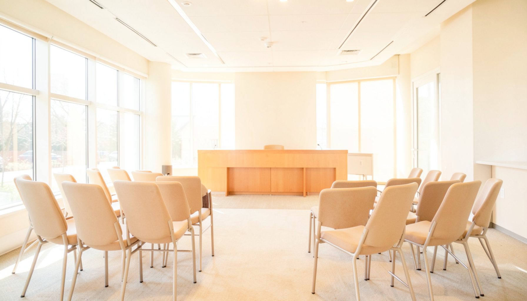 Bright, airy conference room with natural light streaming through large windows, rows of chairs facing a panel table, warm neutral tones