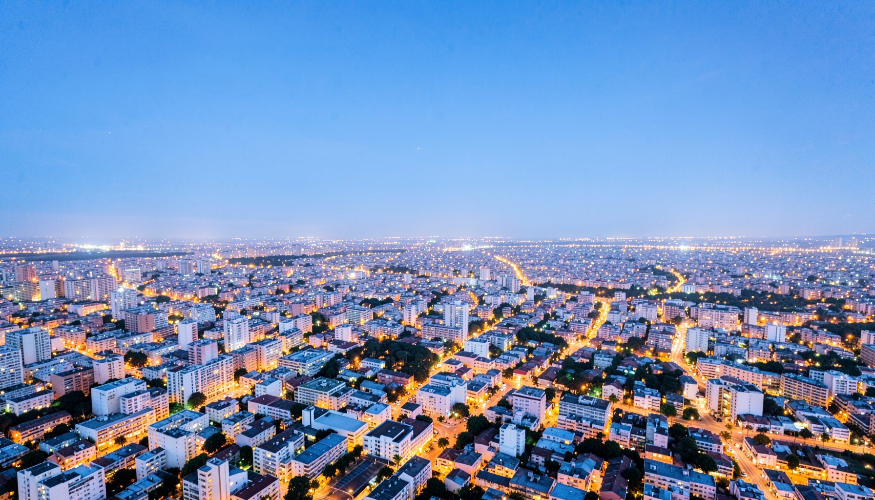Wide aerial view of a sprawling city at dusk with warm amber lights spreading across the urban landscape under a deep blue sky